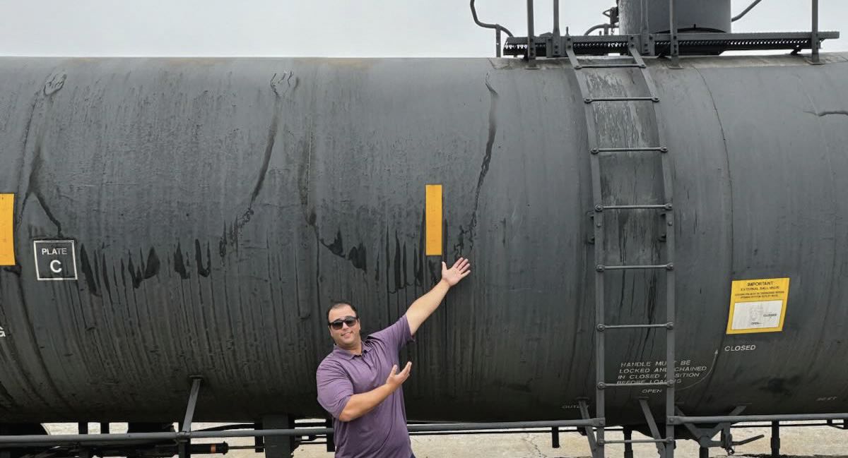 A man in a purple shirt and sunglasses gestures towards a black railcar
