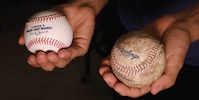 Two hands. One hold a clean baseball and the other holds a dirty one.
