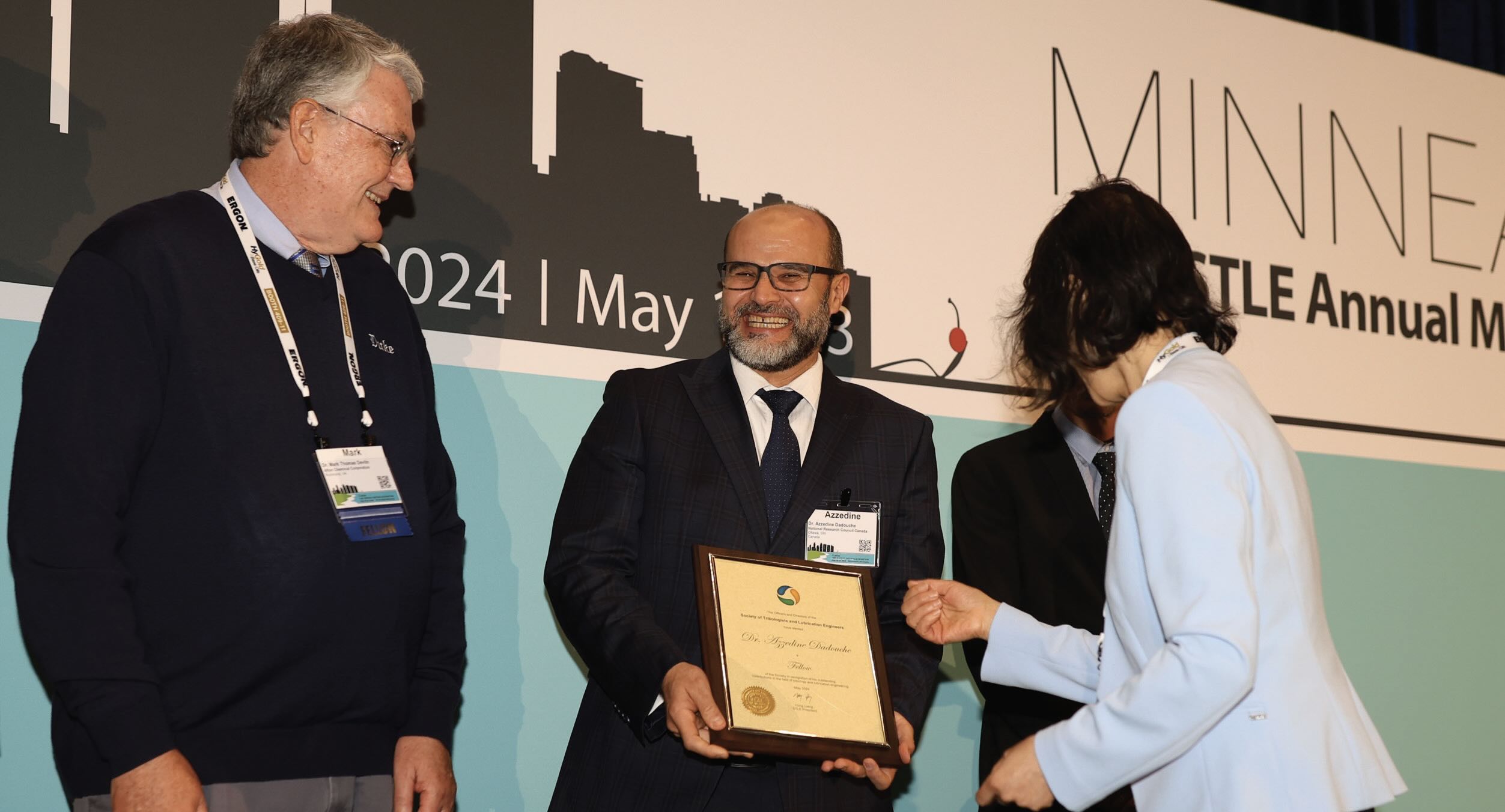 Photo of a woman presenting an award to a smiling man with glasses while another man watches